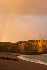 Golden sunset over the cliffs of Étretat, Normandy, France, with a rainbow above the sea. Dramatic coastal landscape showcasing natural beauty and serene atmosphere.
