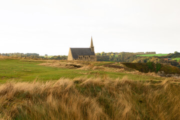 Small chapel on the cliff at Étretat, Normandy, France, surrounded by green hills and cloudy sky. Peaceful coastal landscape with natural and architectural harmony.