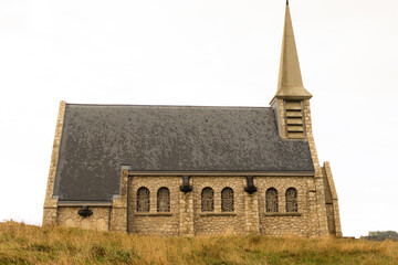 Small chapel on the cliff at Étretat, Normandy, France, surrounded by green hills and cloudy sky. Peaceful coastal landscape with natural and architectural harmony.