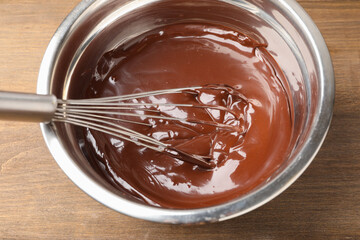Melted chocolate and whisk in bowl on wooden table, closeup