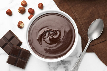 Melted chocolate in bowl, pieces, nuts and spoon on white marble table, flat lay