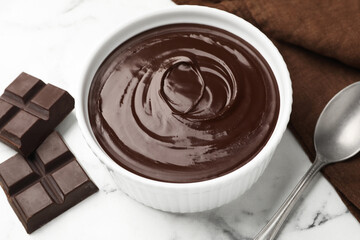 Melted chocolate in bowl, pieces and spoon on white marble table, closeup