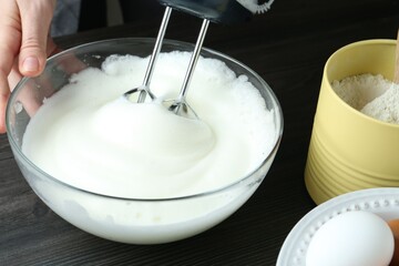 Woman whisking egg whites with hand mixer at black wooden table, closeup