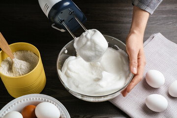 Woman whisking egg whites with hand mixer at black wooden table, closeup