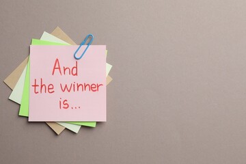 Sticky notes with phrase And The Winner Is on grey background, top view. Space for text