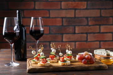 Tasty bruschettas with cheese, cucumber, sausages and red wine on wooden table against brick wall, closeup. Space for text