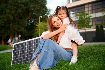 Fototapeta premium Mother and daughter sit together on grass, embracing in warm moment with solar panel beside in the evening. Concept of renewable energy and family, highlighting commitment to sustainable future.