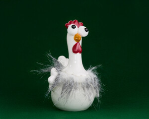 Whimsical white ceramic chicken figurine with googly eyes and fluffy grey feathers, set against a dark green background. A humorous and quirky pet or farm animal representation.