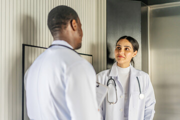 Professional medics wait for elevator in hospital hallway. African American doctor talks about treatment of his patient with Latin Colleague female in medical center corridor. Medical staff at work.