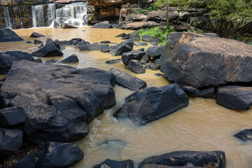 Waterfall flows into a muddy river with large, dark rocks.