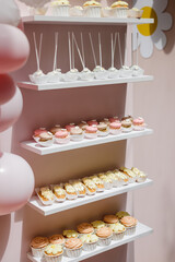 Vertical display of sweet treats, including cupcakes, cake pops, and mini pastries, arranged on white shelves against a pink backdrop, decorated with small flowers and surrounded by balloons