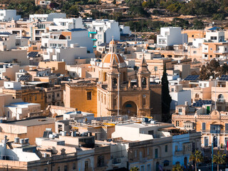 Elevated view of Marsaxlokk, Malta, shows a Baroque church with dome and twin towers amid flat roofs, solar panels, whitewashed blocks, and honey stone homes at midday. © Aerial Film Studio