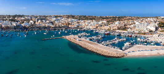 Aerial panorama of Marsaxlokk, Malta, shows the curved breakwater, whitewashed waterfront, marina and quay. Dozens of moored boats rest in turquoise water under clear light. © Aerial Film Studio