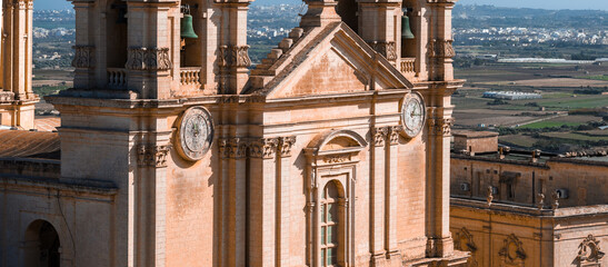 Aerial close up shows St. Paul's Cathedral facade in Mdina, Malta, with twin bell towers, clocks, pilasters, and limestone, looking south to fields and distant towns. © Aerial Film Studio