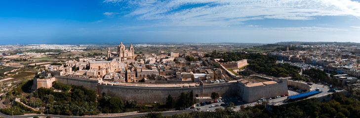Fototapeta premium Aerial panorama of Mdina, Malta, with St. Pauls Cathedral, thick limestone bastion walls, and rural fields under crisp light, contrasting with modern suburbs beyond.