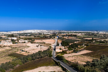 Aerial view over rural outskirts near Mdina, Malta, with patchwork fields, terraced farmland, stone farmhouses, and a road leading to whitewashed urban areas under midday light. © Aerial Film Studio