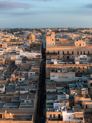 Obraz premium Aerial view of Valletta, Malta, at sunset. A straight street cuts through limestone blocks toward a domed church and fortified palazzi, with deep shadows and warm light.