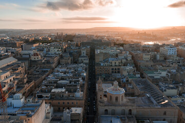 Obraz premium Aerial view looks down Valletta streets at golden hour, domed churches and bastioned walls lead toward Grand Harbour, with shipyards and cranes silhouetted on horizon.