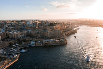 Fototapeta premium Aerial view of Valletta, Malta at sunset, Grand Harbour curving right, boats and a ferry ripple the water, Fort St. Elmo and bastions glow in warm light.