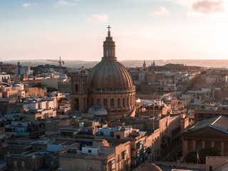 Fototapeta premium Aerial view shows Valletta, Malta at golden hour, the basilica dome and lantern above limestone blocks, long shadows across Baroque streets near Grand Harbour.