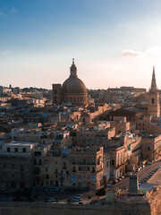 Naklejka premium Aerial view of Valletta, Malta at sunset shows the Basilica dome and St. Pauls Pro Cathedral spire over limestone blocks, warm golden light, long shadows, and a waterfront road.