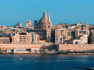 Naklejka premium Valletta, Malta, seen across Grand Harbour, shows the basilica dome, cathedral spire, stone walls, and a small boat in warm late day light with clear blue color.