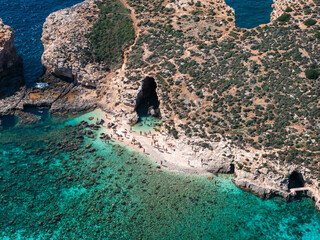 Midday summer aerial shows Comino Island Blue Lagoon, Malta. Turquoise shallows meet rocky beach, limestone cliffs with two sea caves, swimmers by the larger cavern.