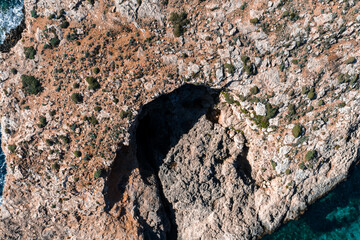 Aerial top down view of Comino Island, Malta, shows a sea cave by Blue Lagoon. Sunlit limestone, low shrubs, and jagged fracture lines appear in bright midday light.