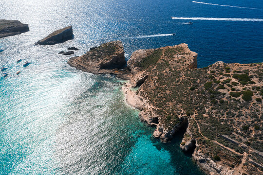 Aerial midday view of Blue Lagoon on Comino, Malta, with Cominotto channel, speedboat wakes, turquoise to deep blue gradient, rocky coves, sandy inlet, and arid cliffs.