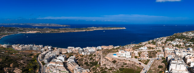 Aerial view shows Mellieha on Malta north coast, terraced white buildings toward Mellieha Bay, Comino and Gozo on horizon, midday light, clear view, small boats © Aerial Film Studio