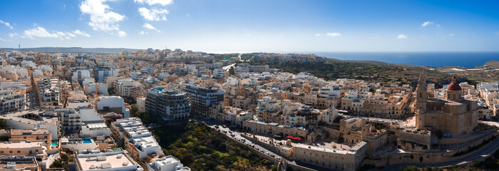 Aerial view shows Mellieha, Malta, terraced hills, limestone blocks, and twin spire parish church. Streets lead to coast, Comino and Gozo appear in clear midday light. © Aerial Film Studio