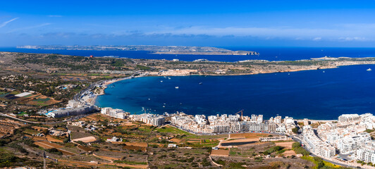Aerial panorama of Mellieha Bay in Malta shows a crescent beach, whitewashed buildings, and cobalt water. Comino and Gozo line the horizon, boats dot the channel. © Aerial Film Studio