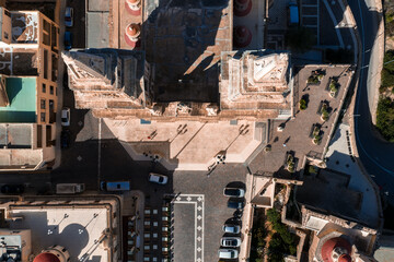 Aerial top view shows the sanctuary forecourt, twin bell towers, and stone plaza in Mellieha, Malta. Warm light reveals honey limestone, red domes, cars, and curving streets. © Aerial Film Studio