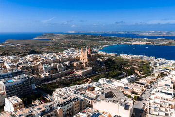 Aerial panorama of Mellieha, Malta shows Mellieha Parish Church above limestone buildings, Mellieha Bay, and Comino and Gozo on the horizon in bright daylight.