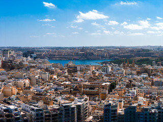 Aerial view shows Sliema, Malta, with flat roofs and modern blocks leading to Marsamxett Harbour. Valletta domes and bastions appear across the water in midday light.