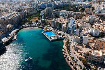 Fototapeta premium Aerial view of Sliema, Malta shows a turquoise inlet, a seaside lido with a large rectangular pool on a pier, curving apartments, a promenade, light traffic, and a drifting boat.