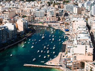 Aerial panorama of a narrow turquoise inlet in Sliema near Spinola Bay, Malta. Dozens of small white boats sit moored by a curving promenade and mid rise apartments at midday. © Aerial Film Studio