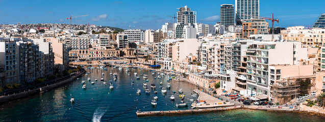 Fototapeta premium Aerial view shows St. Julian's and Sliema harbor in Malta with mid rise apartments, towers, moored boats, curving bay, clear daylight, and cranes signaling development.