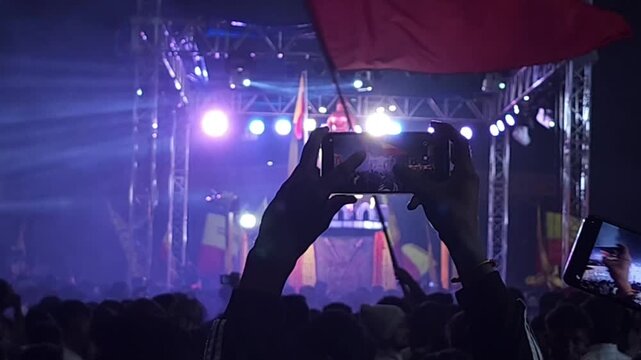 A massive crowd gathers at night for a public cultural celebration in India. People watch performances on a decorated stage with bright lights, flags, and banners
