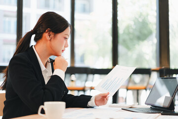 Young businesswoman reviewing financial report at bright office table, focused expression, coffee cup nearby