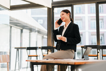 Young businesswoman talking on phone while holding coffee cup in modern bright coworking space, confident and relaxed