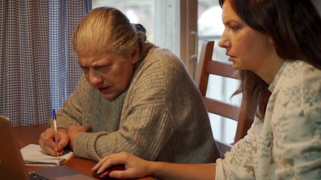 An elderly woman is holding a pen and concentrating intently on a laptop screen while a younger woman operates the computer mouse. They are working together at a wooden table