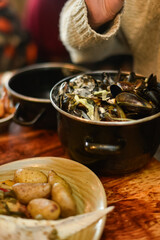 A vertical close-up of a pot filled with freshly cooked mussels, next to a shallow bowl of roasted potatoes and vegetables. Focuses on the texture of the shellfish and rustic meal presentation. 