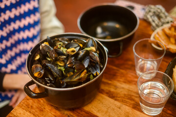 A pot of hot, freshly cooked mussels in a sauce sits on a wooden table, next to two glasses of water or wine. Represents traditional French cuisine and a warm, shared dining experience. Horizontal.