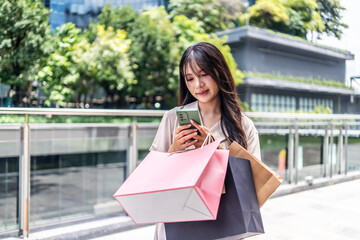 Young asian woman Stand and type a message on your mobile phone. at a lively shopping area, holding her bags with excitement. Her bright smile reflects the joy of a successful shopping trip.
