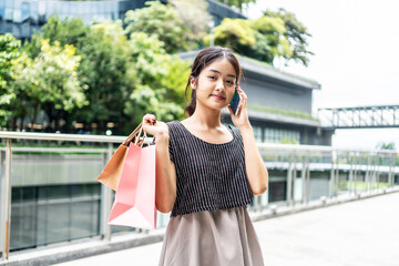 Young Asian woman shopping with paper bags walking in the city while holding a smartphone with happiness. Happy lady shopping outdoors in a department store, Smiling girl enjoying lifestyle fashion