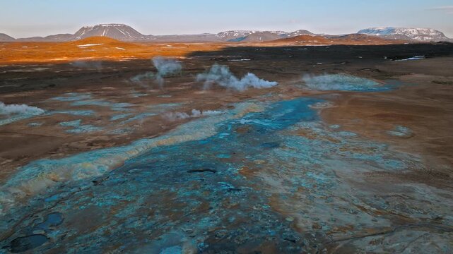 Drone shot of Hverir geothermal area near Lake Mývatn, North Iceland. Steam rises from fumaroles across blue and orange volcanic soil.