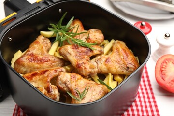 Grilled chicken wings with french fries and rosemary in air fryer basket on white wooden table, closeup