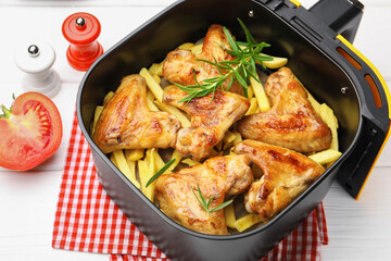 Grilled chicken wings with french fries in air fryer basket, tomato and tongs on white wooden table, closeup