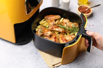 Woman opening air fryer with grilled chicken wings and french fries at grey table, closeup